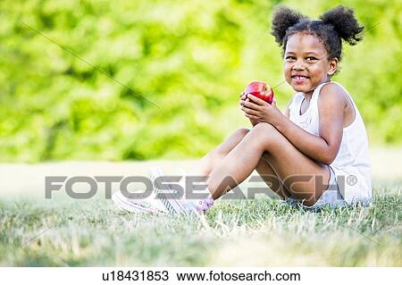 Stock Image - Girl sitting and holding apple in park.. Fotosearch