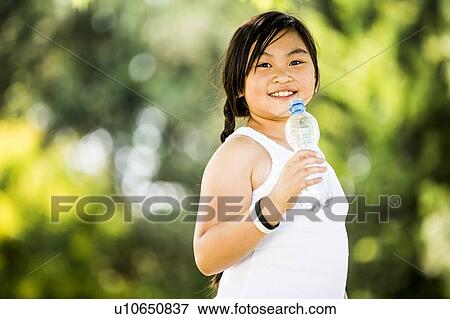 Stock Photo - Girl standing with water bottle in park, smiling, portrait.. Fotosearch