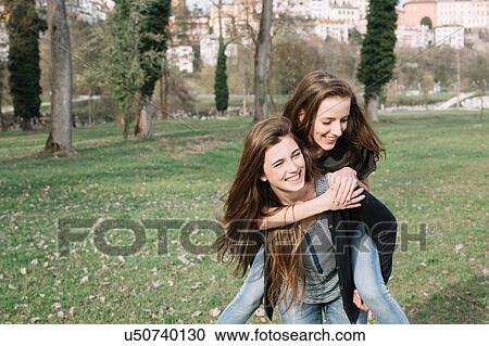Stock Image - Girlfriends playing piggyback ride in park. Fotosearch