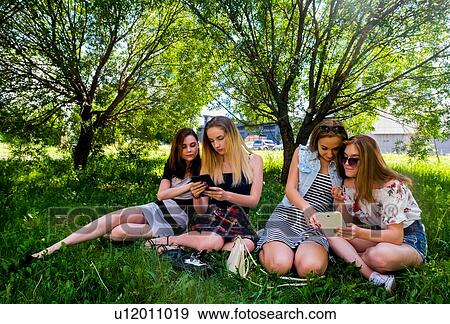 Stock Photo - Girlfriends relaxing, using smart devices in city park. Fotosearch