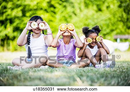 Stock Image - Girls sitting side by side in park and covering their eyes with fruit slices, smiling.. Fotosearch