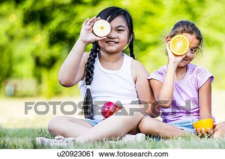 Stock Image - Girls sitting side by side in park and holding various fruits, smiling, portrait.. Fotosearch