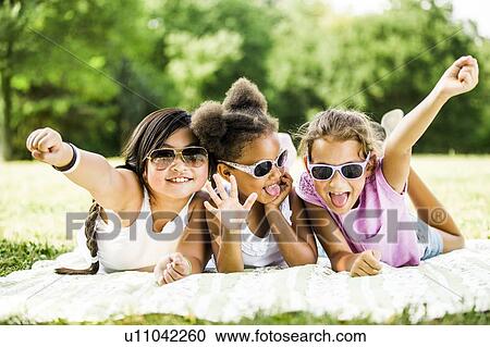 Stock Image - Girls wearing sunglasses and lying side by side on blanket in park, smiling, portrait.. Fotosearch