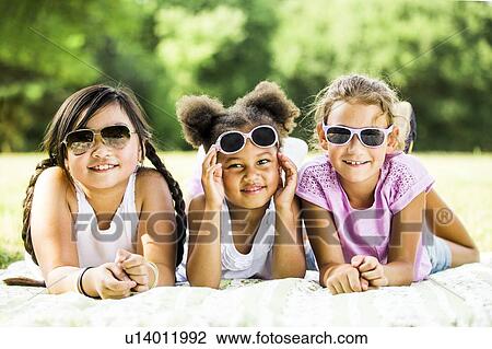 Stock Image - Girls wearing sunglasses and lying side by side on blanket in park, smiling, portrait.. Fotosearch