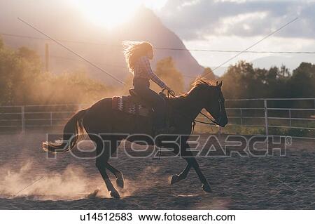 Stock Image - Long haired young woman cantering on horse in rural equestrian arena, backlit, Primaluna, Trentino-Alto Adige, Italy. Fotosearch