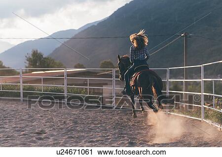 Stock Image - Long haired young woman cantering on horse in rural equestrian arena, rear view, Primaluna, Trentino-Alto Adige, Italy. Fotosearch