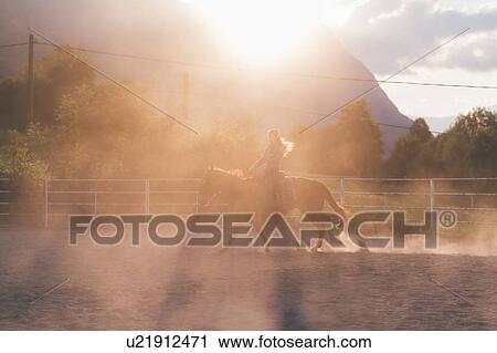 Stock Image - Long haired young woman trotting on horse in rural equestrian arena, backlit, Primaluna, Trentino-Alto Adige, Italy. Fotosearch