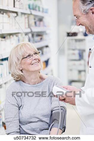 Stock Image - Male pharmacist checking smiling senior female patient's blood pressure in pharmacy.. Fotosearch