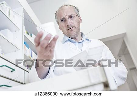 Stock Image - Male pharmacist reading label on pill bottle while looking for medicine in drawer cabinet in pharmacy.. Fotosearch