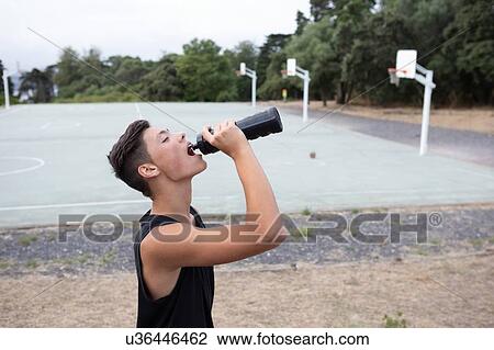 Stock Image - Male teenage basketball player drinking from water bottle on basketball court. Fotosearch
