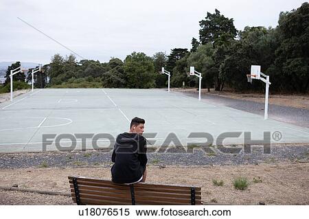 Stock Photography - Male teenage basketball player looking back from park bench by basketball court. Fotosearch