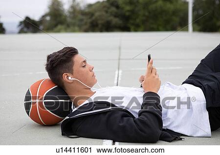 Stock Image - Male teenage basketball player lying on basketball court looking at smartphone. Fotosearch