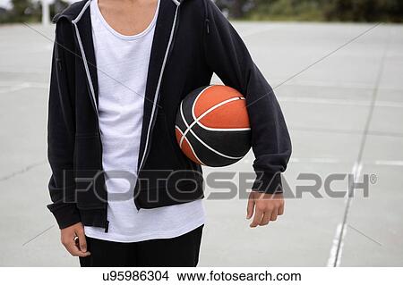 Picture - Male teenage basketball player on basketball court with ball under his arm, mid section. Fotosearch