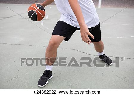 Stock Image - Male teenage basketball player playing basketball on court, waist down. Fotosearch