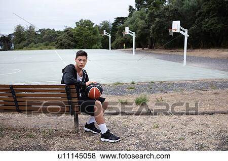 Stock Photo - Male teenage basketball player sitting on park bench by basketball court, portrait. Fotosearch