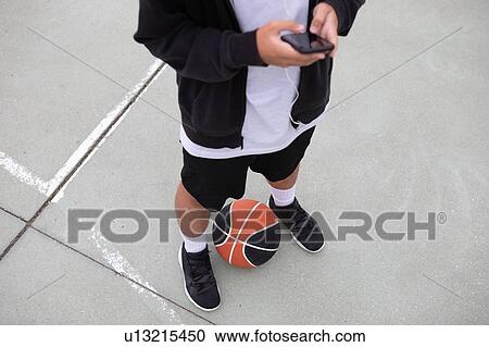 Stock Image - Male teenage basketball player standing on basketball court looking at smartphone, waist down. Fotosearch