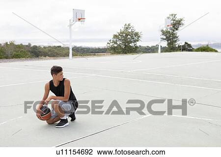 Stock Image - Male teenage basketball player crouching with ball on basketball court. Fotosearch