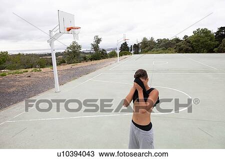 Stock Image - Male teenage basketball player wiping his brow with vest on basketball court. Fotosearch