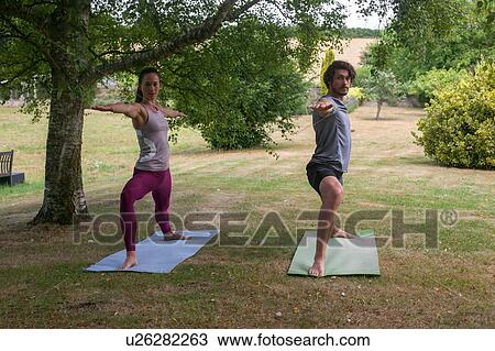 Stock Image - Man and woman doing yoga in garden, practicing warrior pose. Fotosearch