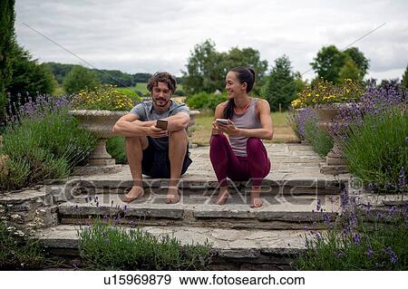 Stock Photo - Man and woman practicing yoga in garden, looking at smartphones on patio. Fotosearch