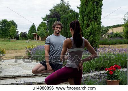 Stock Photograph - Man and woman practicing yoga in garden, tree pose. Fotosearch