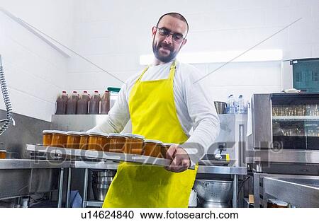 Stock Image - Man carrying tray of fresh jam in industrial kitchen. Fotosearch