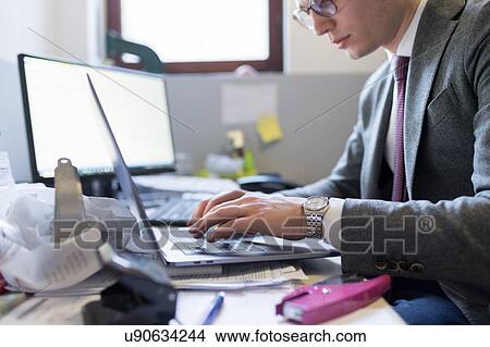 Picture - Man using laptop and desktop in office. Fotosearch
