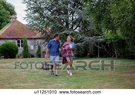 Stock Image - Mature couple strolling across garden lawn with picnic blanket. Fotosearch