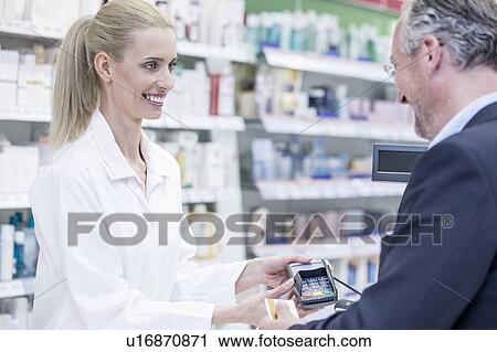 Stock Image - Mature man paying female pharmacist in pharmacy.. Fotosearch