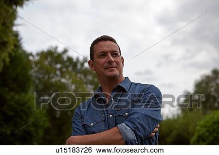 Stock Photograph - Mature man with arms crossed in garden, head and shoulder portrait. Fotosearch