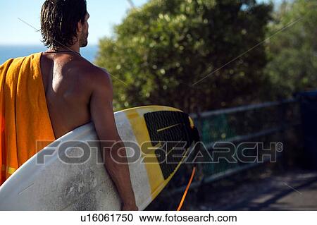 Stock Image - Mid adult male surfer carrying surfboard along beach walkway, rear view, Camps Bay Beach, Cape Town, South Africa. Fotosearch
