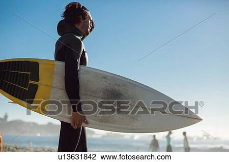 Stock Image - Mid adult male surfer in wet suit carrying surfboard on beach, Camps Bay Beach, Cape Town, South Africa. Fotosearch