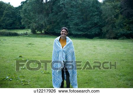 Stock Photograph - Mid adult woman standing in rural field wrapped in blanket, full length portrait. Fotosearch