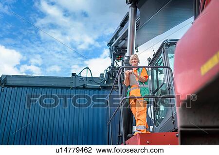 Stock Image - Portrait of female container lift operator with shipping container in port. Fotosearch