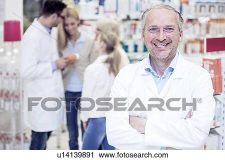 Stock Image - Portrait of mature male pharmacist standing in pharmacy, with colleague and customers in the background.. Fotosearch
