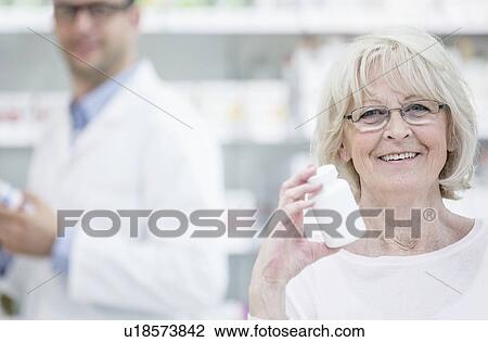 Stock Image - Portrait of senior woman holding pill bottle in pharmacy, with pharmacist in the background.. Fotosearch