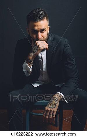 Stock Image - Portrait of young man, sitting on chair, wearing suit, tattoos on hands, pensive expression. Fotosearch
