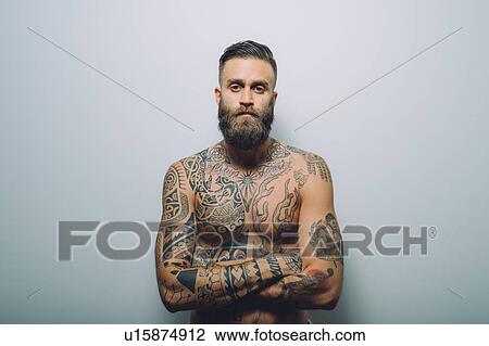 Stock Image - Portrait of young man with beard, bare chest covered in tattoos, arms folded. Fotosearch