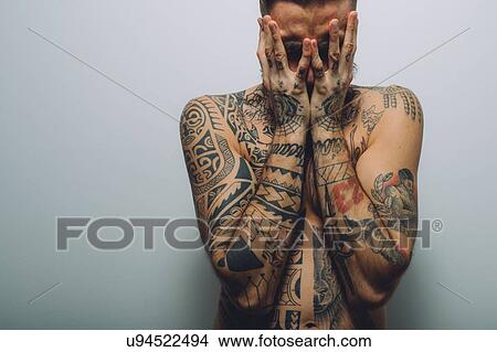 Picture - Portrait of young man with beard, covered in tattoos, hands covering face. Fotosearch