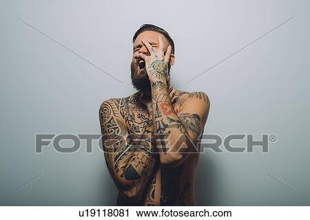 Stock Image - Portrait of young man with beard, covered in tattoos, hand on face, screaming. Fotosearch