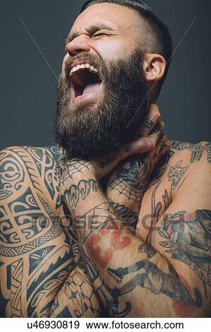 Stock Photo - Portrait of young man with beard, covered in tattoos, hands around throat, screaming. Fotosearch