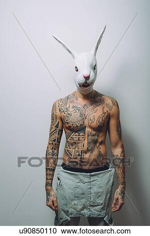 Stock Image - Portrait of young man with wearing rabbit mask, bare chest covered in tattoos. Fotosearch