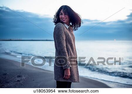 Picture - Portrait of young woman standing on beach, looking over shoulder, smiling. Fotosearch