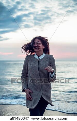 Portrait of young woman standing on beach, her back to the ocean, wind blowing her hair, smiling View Large Photo Image Stock Photo - Portrait of young woman standing on beach, her back to the ocean, wind blowing her hair, smiling. Fotosearch