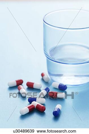 Stock Photo - Red and blue drug capsules scattered on a blue background with a glass of water beside them.. Fotosearch