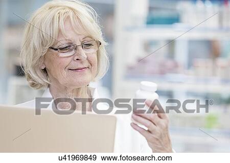 Stock Photo - Senior female pharmacist holding laptop and reading label on pill bottle in pharmacy.. Fotosearch