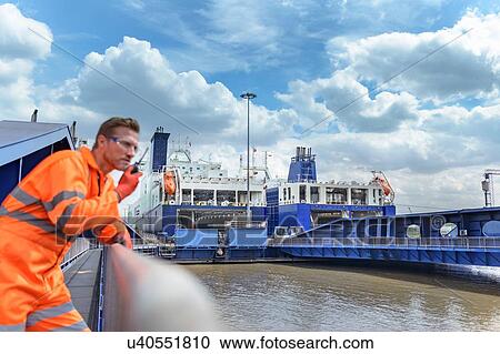 Stock Image - Ship's worker using walkie talkie to communicate with loading ferry ships in port in background. Fotosearch