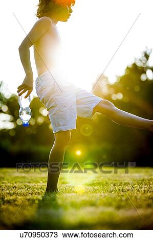 Stock Image - Shirtless boy stretching his leg in park with a water bottle in his hand, backlit.. Fotosearch