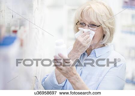 Stock Photo - Sick senior woman reading label on pill bottle in pharmacy while blowing nose.. Fotosearch
