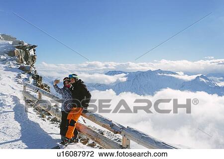 Stock Image - Skiing couple taking smartphone selfie above the clouds, Madonna di Senales, Trentino-Alto Adige, Italy. Fotosearch